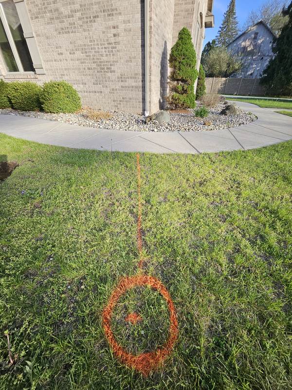 Orange spray-paint line marked across a front lawn from a downspout at a brick house, ending in a painted circle where the pop-up emitter will go